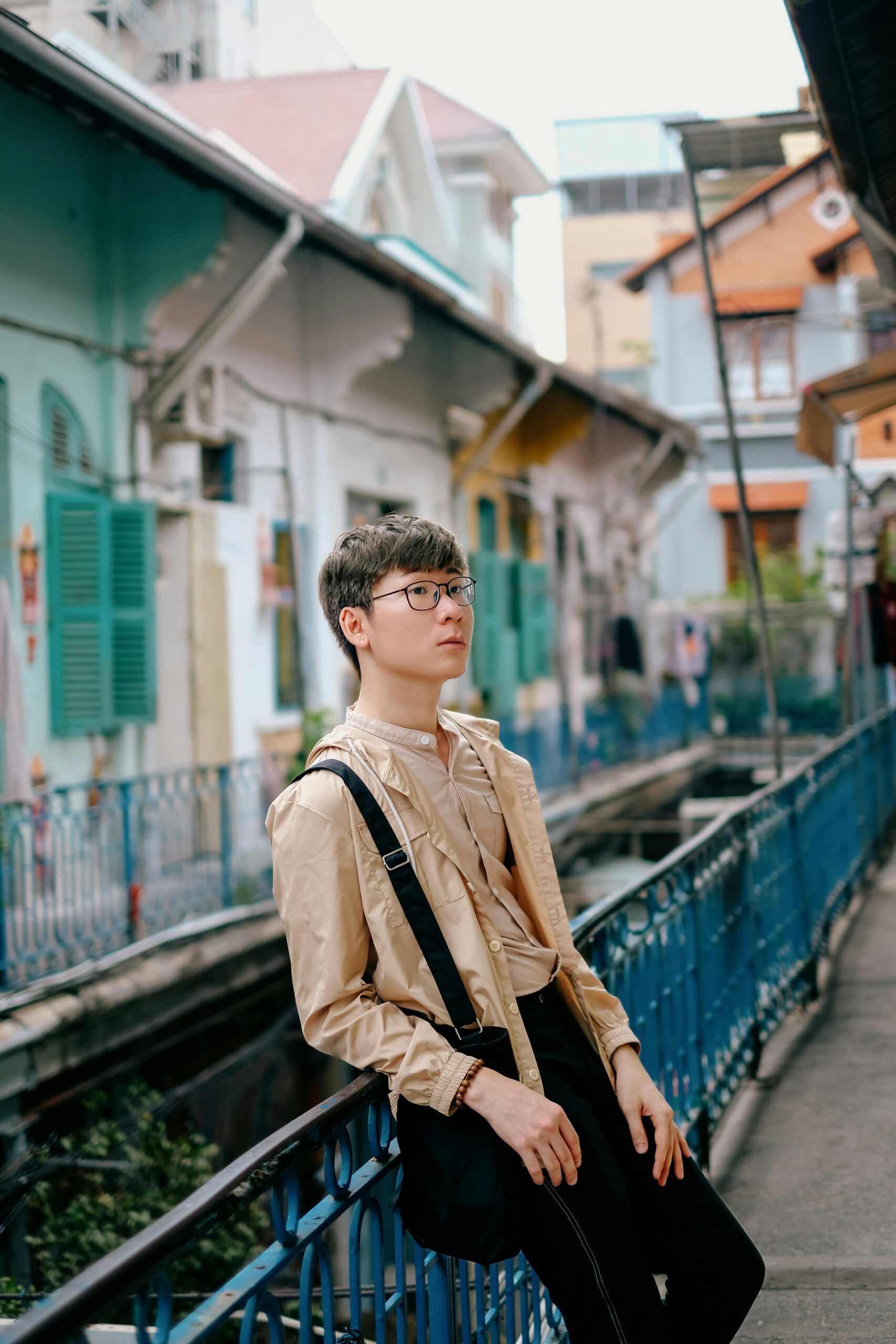 A fashionable young man stands against a railing in a vibrant urban setting, exuding modern style and relaxed vibes.