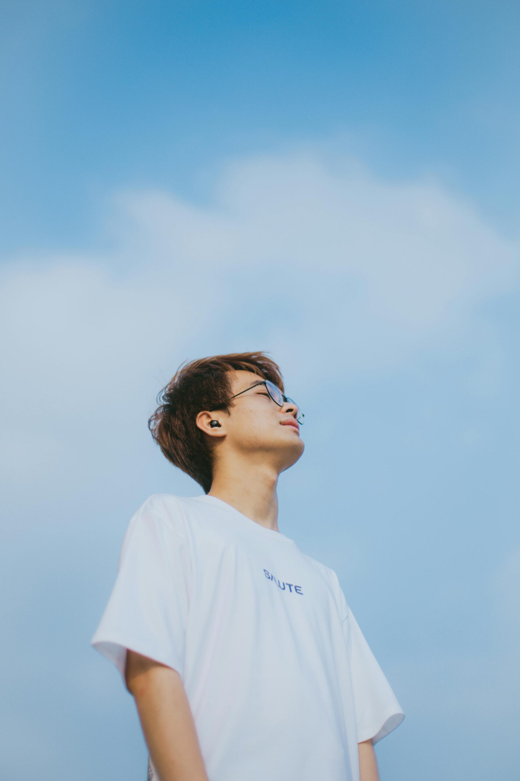 A serene portrait of a young Asian man in a white t-shirt gazing up, set against a bright blue sky.